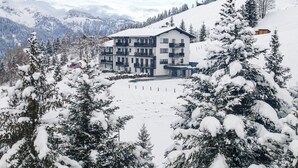 Um edifício coberto de neve com vários andares, rodeado por árvores carregadas de neve e montanhas ao fundo.