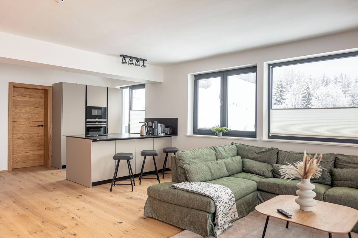 Eine moderne Küche mit Kochinsel, Barhocker und einem grünen Sofa in einem Wohnbereich mit großen Fenstern.