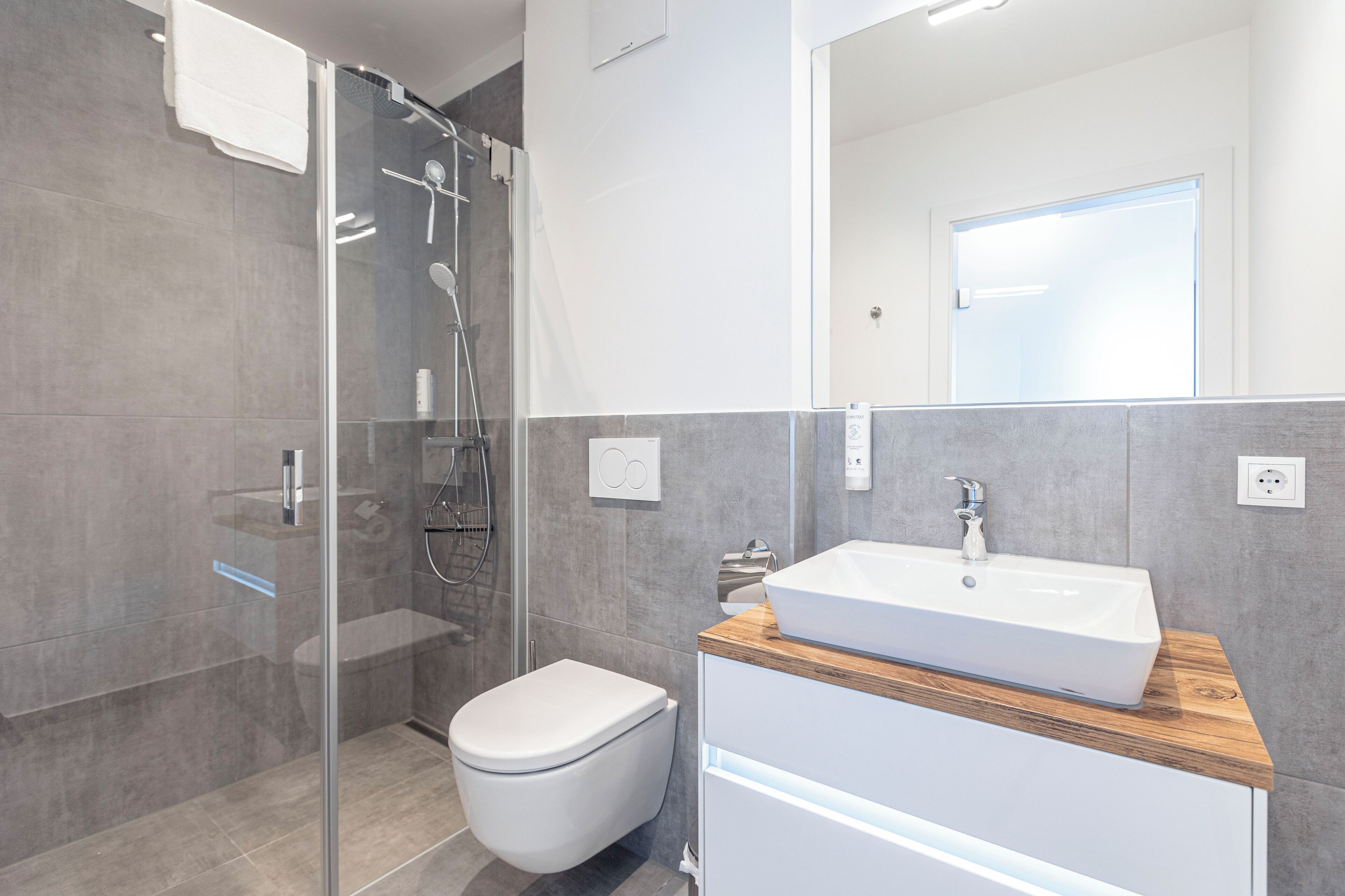 Ein modernes Badezimmer mit einer gläsernen Duschkabine, einer weißen Toilette und einem hölzernen Waschtisch mit weißem Waschbecken.