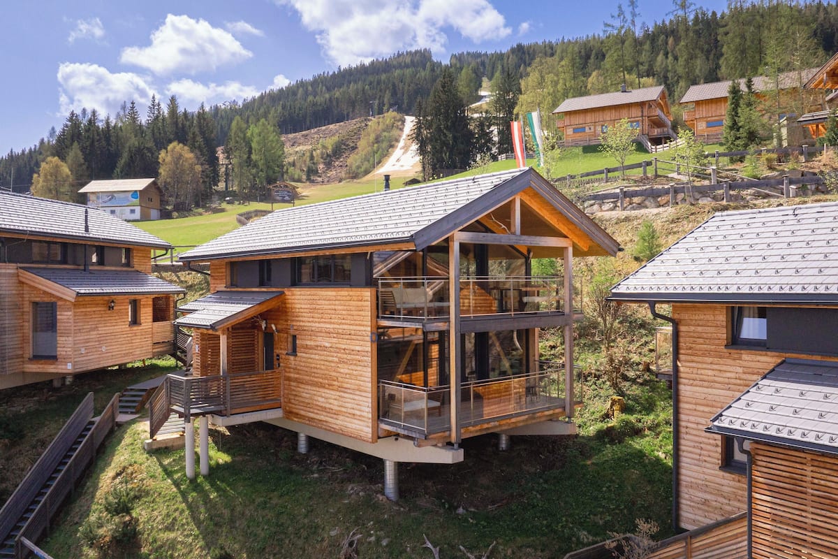 A cluster of wooden cabins with a glass balcony, set in a mountainous area with a clear sky.