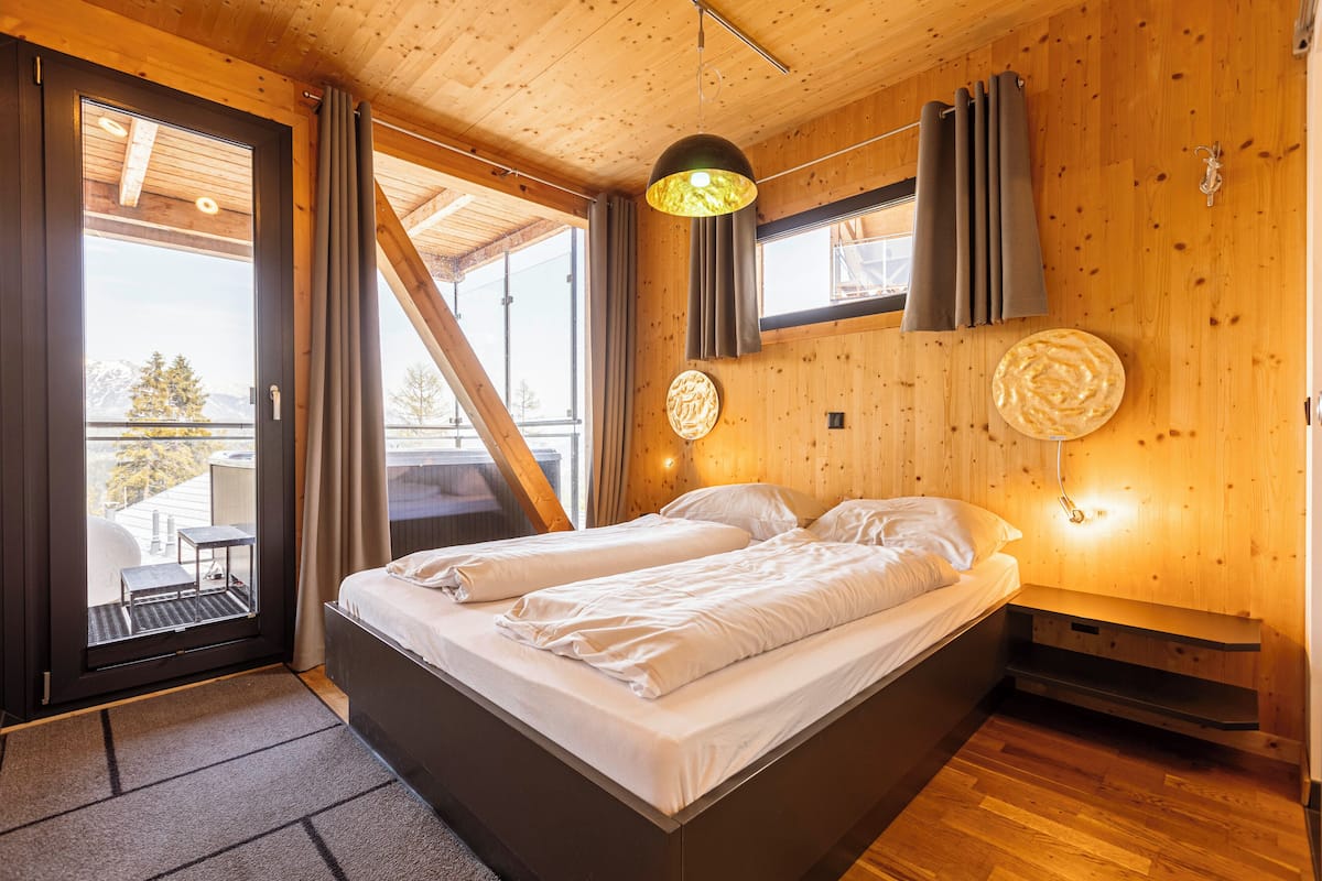 A wooden cabin bedroom with a bed, a small table, and a view of the outdoors.