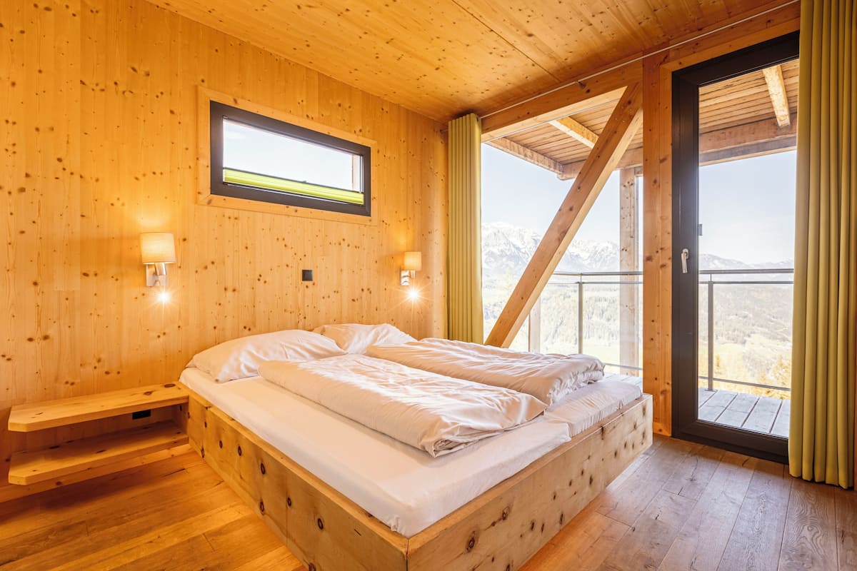 A wooden room with a bed, a window, and a view of mountains.