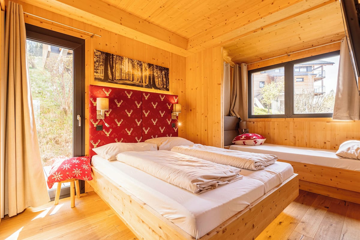 A wooden cabin bedroom with a large bed, a bench, and a window with a view of buildings.