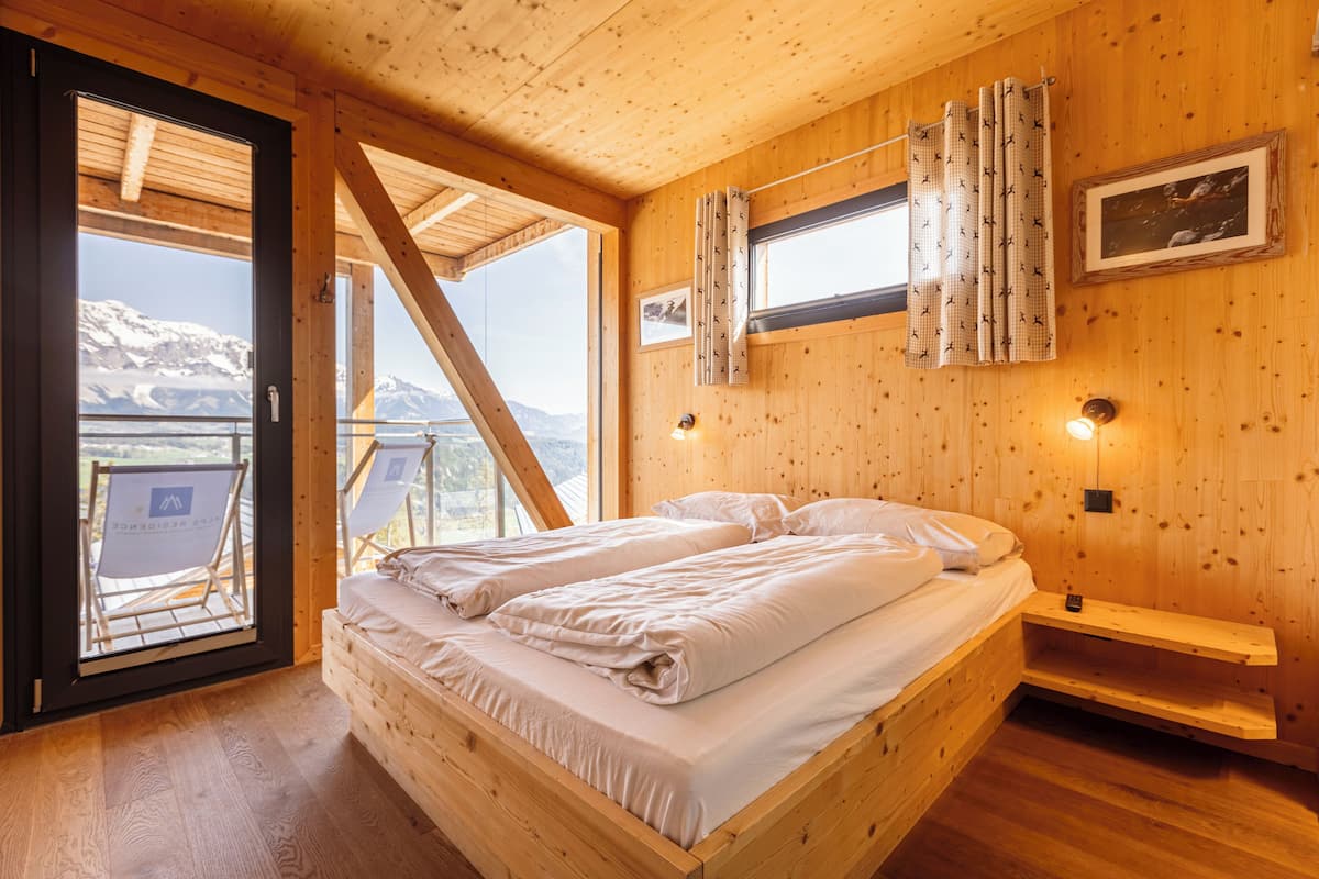 A wooden cabin bedroom with a bed, a small bedside table, a window with curtains, and a view of mountains.