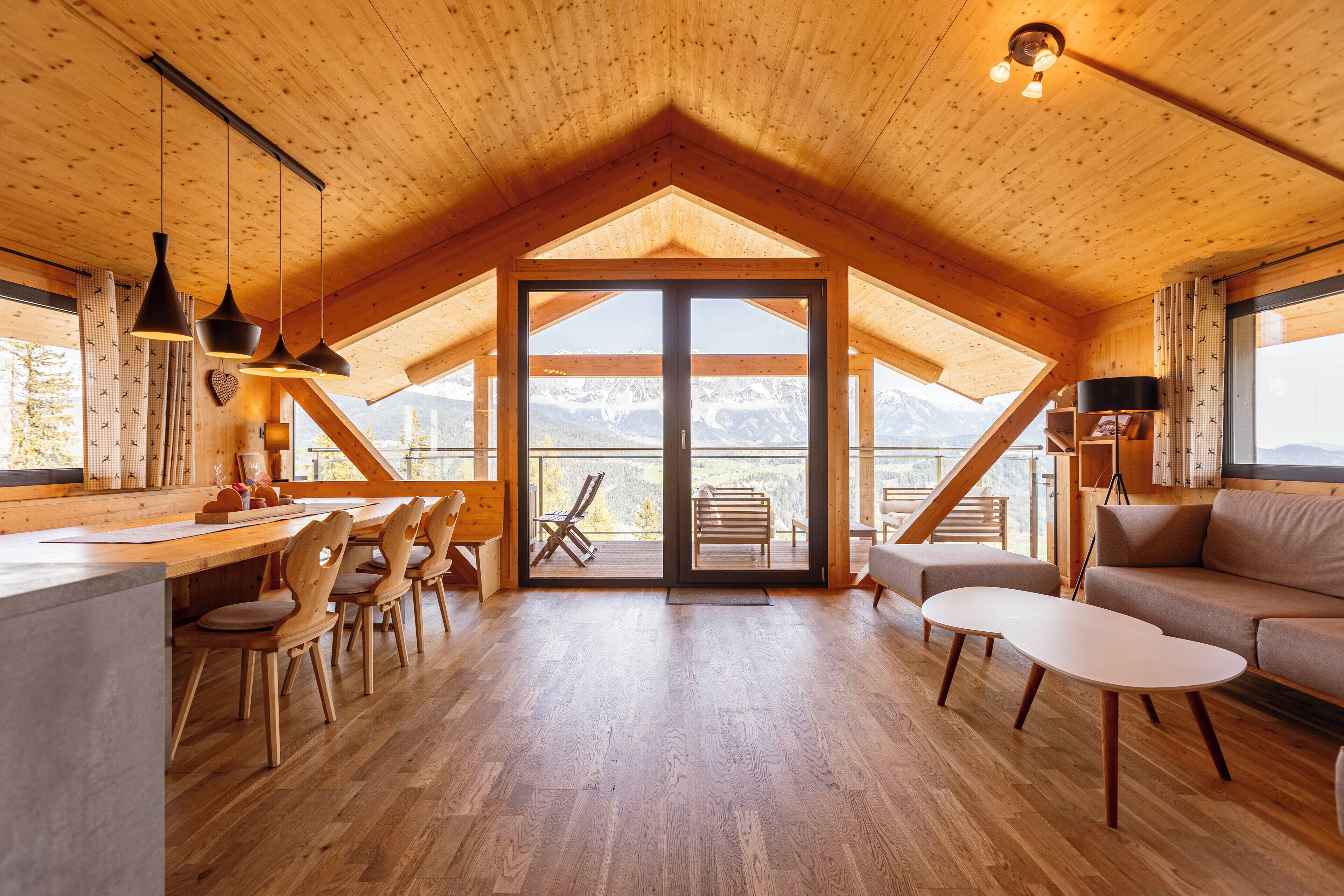 Un vaste espace de vie de style loft, doté de planchers en bois et d’un plafond en pente, comprenant un coin repas avec des chaises et une table, un canapé et une grande porte-fenêtre coulissante donnant sur un balcon avec vue sur les montagnes.