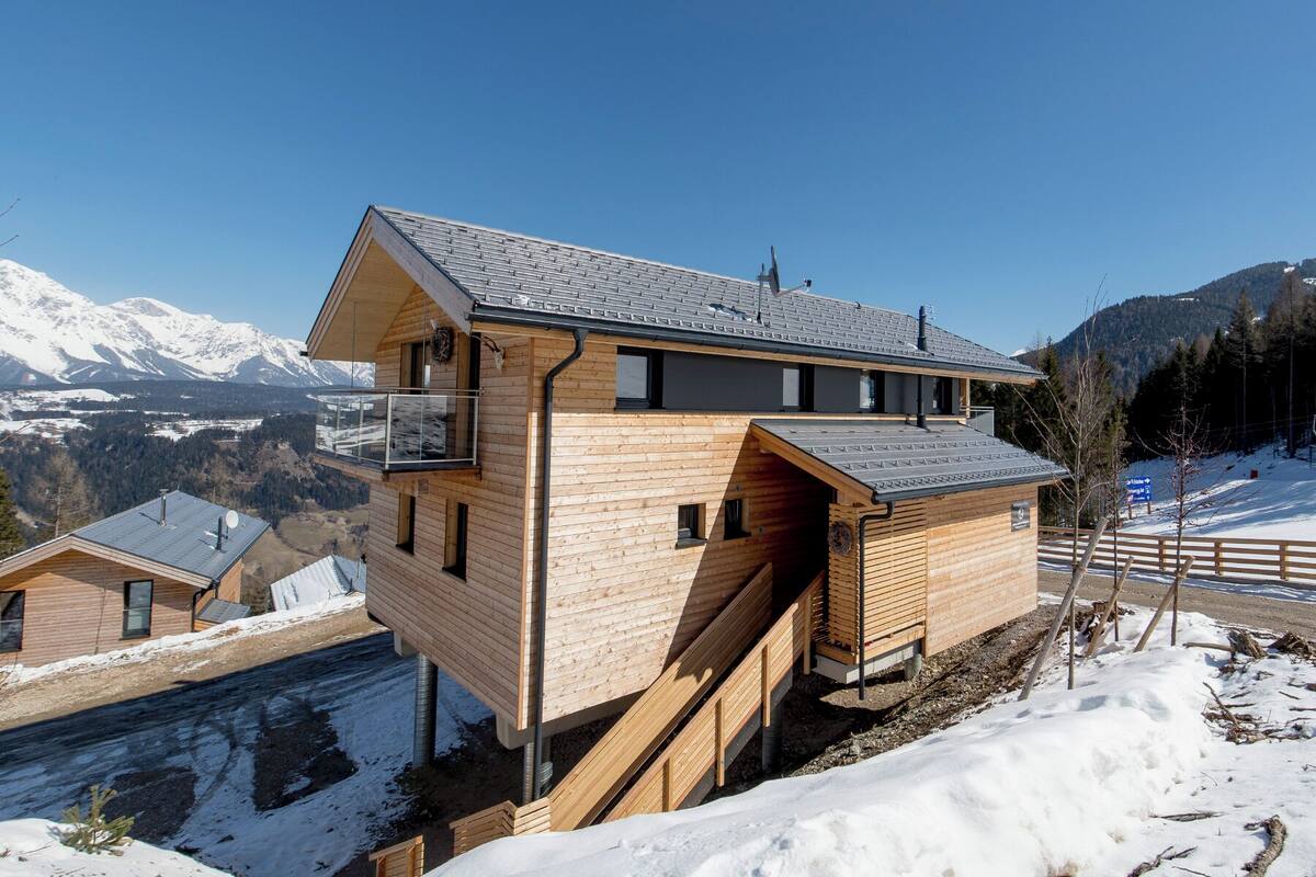 A wooden chalet with a balcony, a staircase, and a view of snow-covered mountains.
