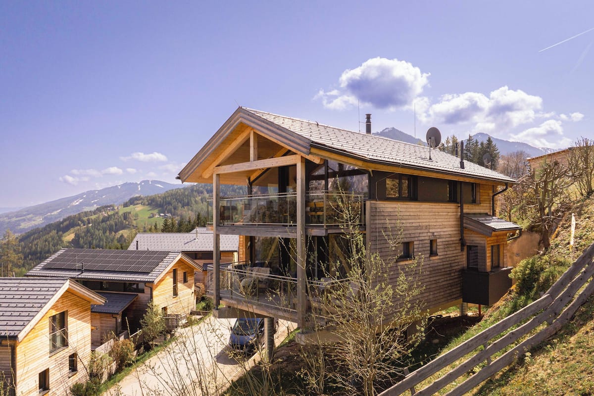 A modern wooden house with a large balcony, solar panels, and a view of mountains.