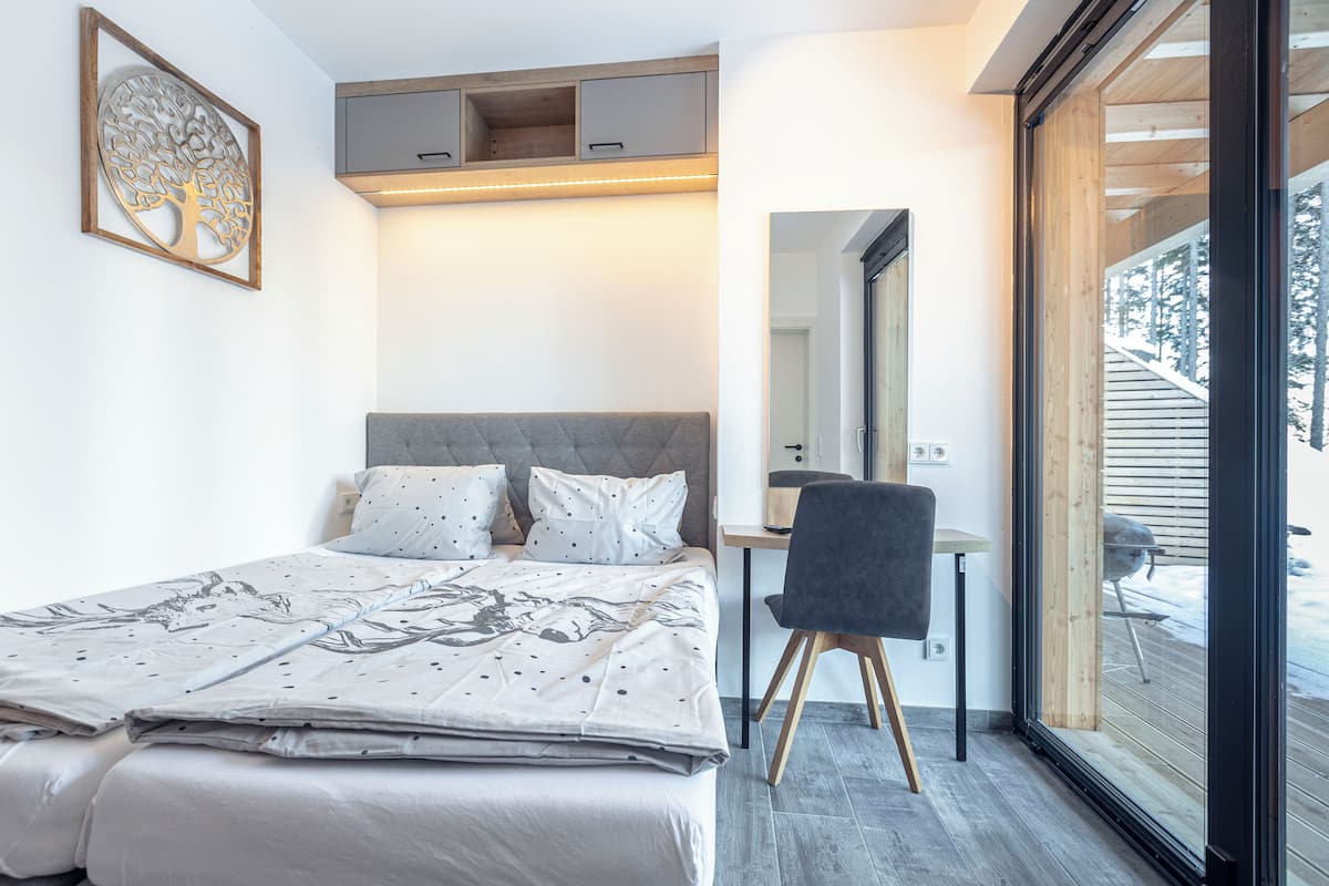 A bedroom with a bed, a desk, a chair, and a large window with a view of a wooden deck.