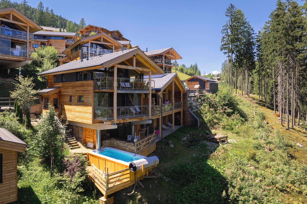 A multi-story wooden chalet with balconies, a hot tub, and a pool, surrounded by forested hills.