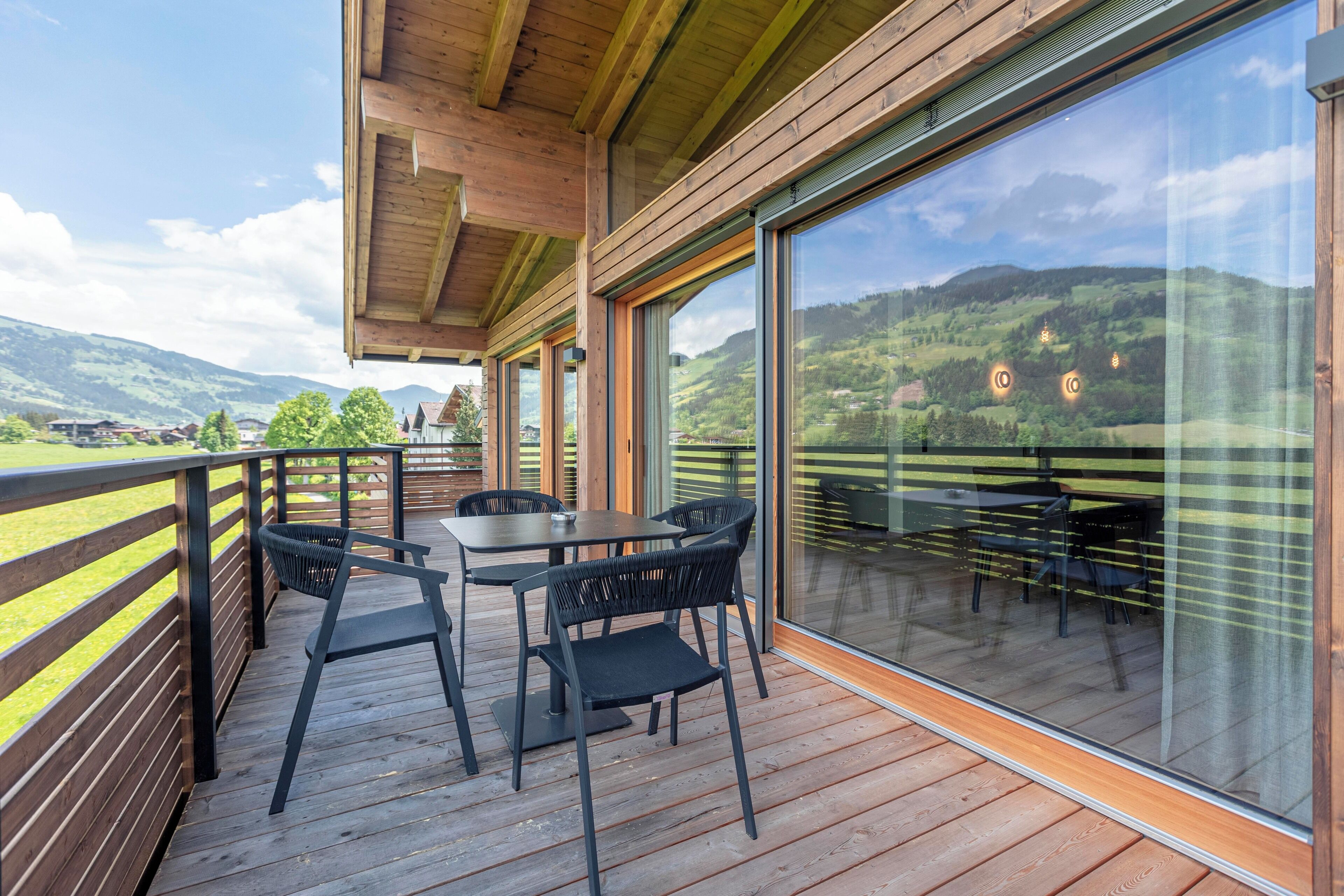 Eine Holzterrasse mit Sitzgelegenheiten im Freien und großen Fenstern, die einen Blick auf eine grüne Landschaft bieten.