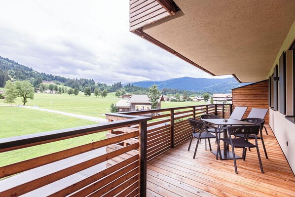 A wooden deck with a table and chairs, overlooking a green field and mountains.