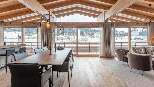 A spacious dining area with a long wooden table and chairs, large windows offering a view of a snowy landscape, and a cozy seating area with a sofa and armchairs. - Chalet 149 Westendorf by ALPS RESORTS (Westendorf)