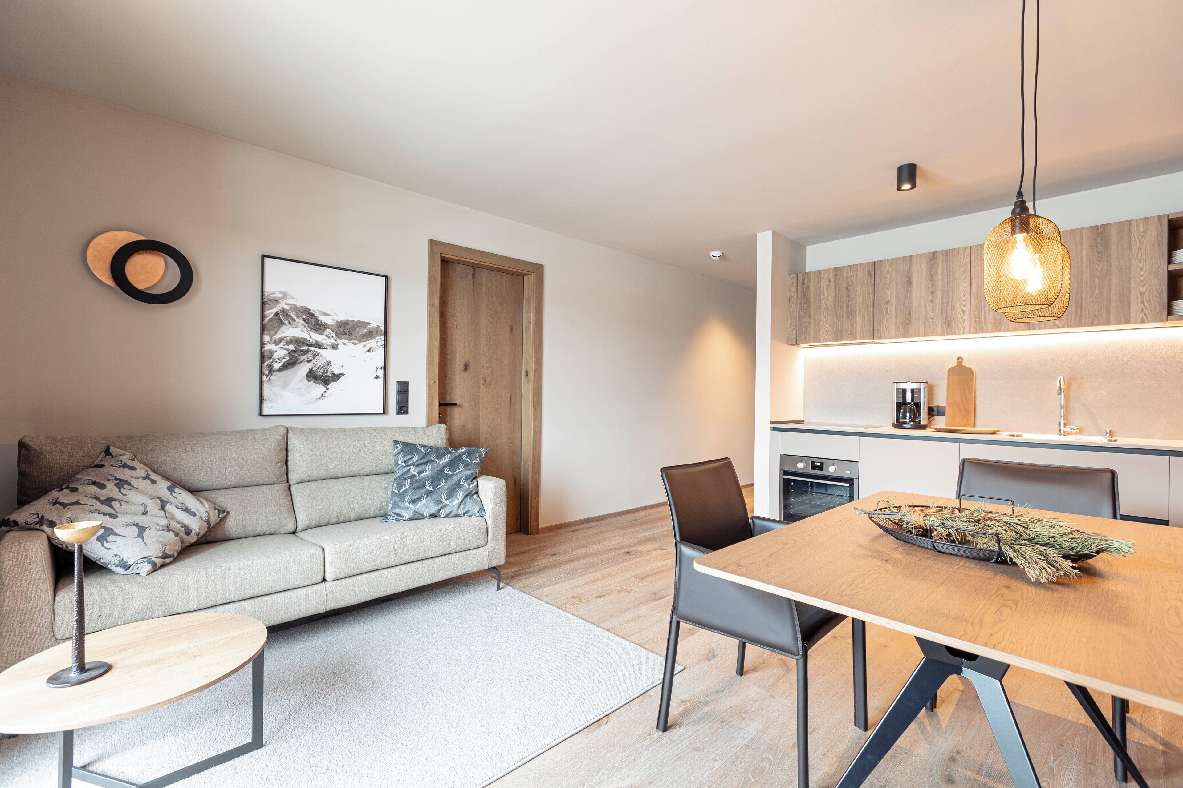 Una sala de estar moderna con sofá, una mesita de centro y una mesa de comedor. Hay una zona de cocina con armarios de madera y una lámpara colgante.