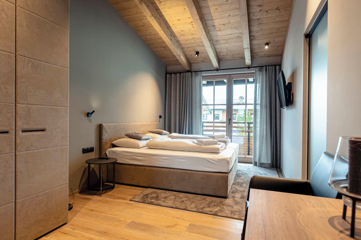 Ein modernes Schlafzimmer mit Holzboden, einem großen Bett mit Kissen, einem kleinen Nachttisch und Blick ins Freie durch ein Fenster.