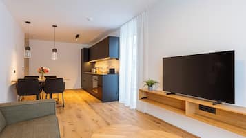 Una sala de estar moderna con un televisor de pantalla plana montado en un estante de madera, una zona de comedor con mesa y sillas, y una cocina al fondo.