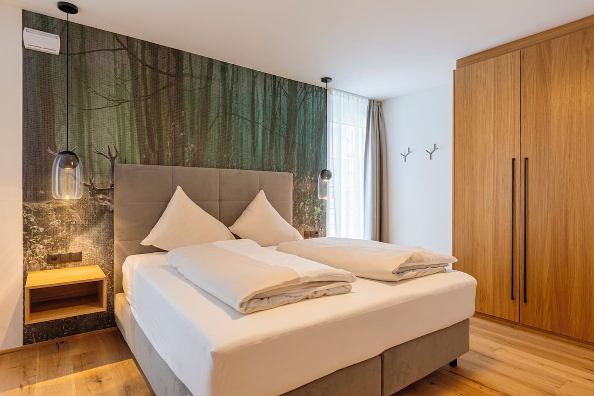 A bedroom with a bed, a nightstand, a wardrobe, and a wall with a forest mural.