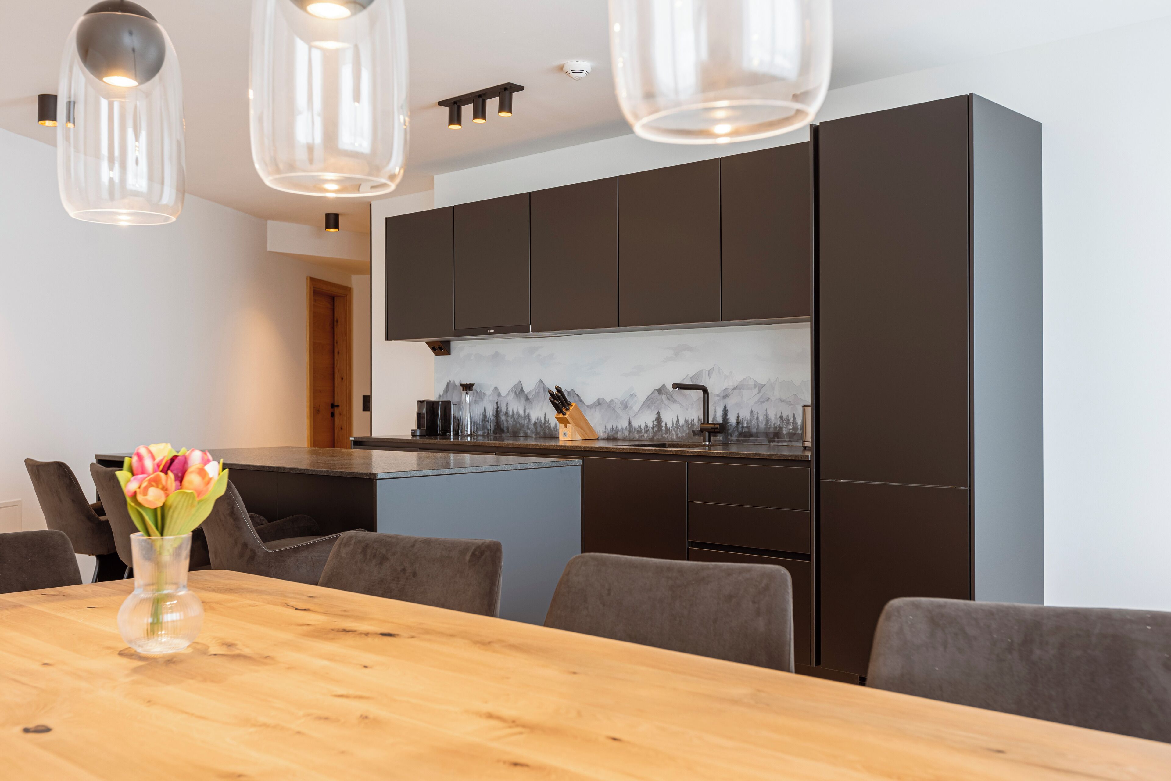 A modern kitchen with dark cabinets, a wooden dining table, and a vase of flowers.