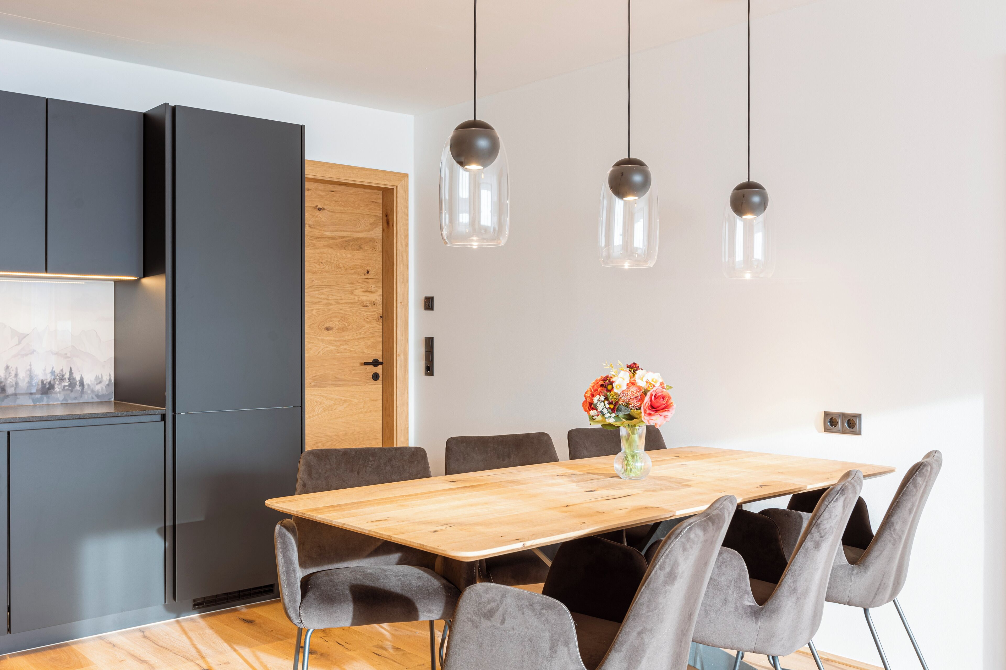 Un espace de salle à manger moderne avec une table en bois, des chaises à revêtement gris et des luminaires suspendus.