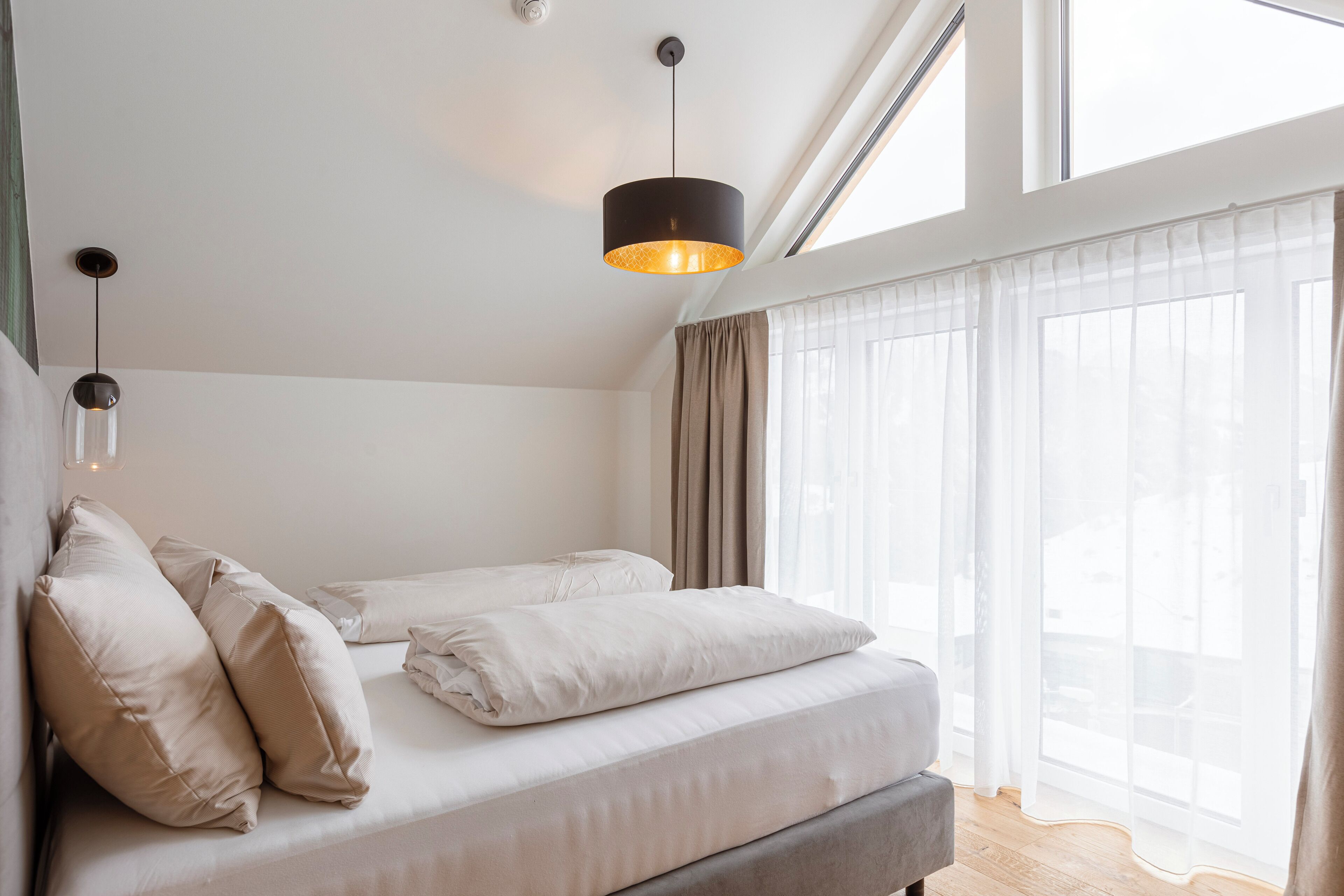 A modern bedroom with a bed, two bedside lamps, and a large window with sheer curtains.