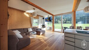 A modern living room with a wooden ceiling, a large sofa, a TV, and a view of a green landscape through large windows. - Feriendorf Murau by ALPS RESORTS (Murau)