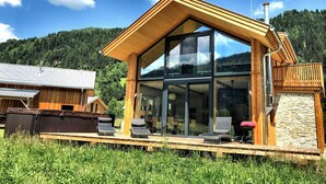A modern wooden cabin with large glass windows, a deck with outdoor seating, and a forested backdrop. - Feriendorf Murau by ALPS RESORTS (Murau)