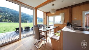 A modern kitchen with a large island, wooden beams, and a view of a green landscape. - Feriendorf Murau by ALPS RESORTS (Murau)