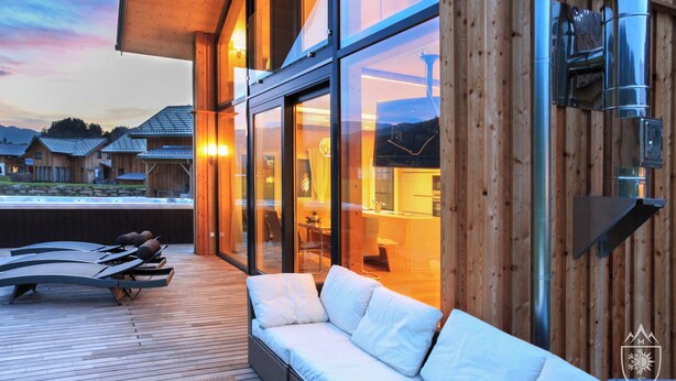 A modern outdoor lounge area with a wooden deck, a white sofa, and lounge chairs. - Feriendorf Murau by ALPS RESORTS (Murau)