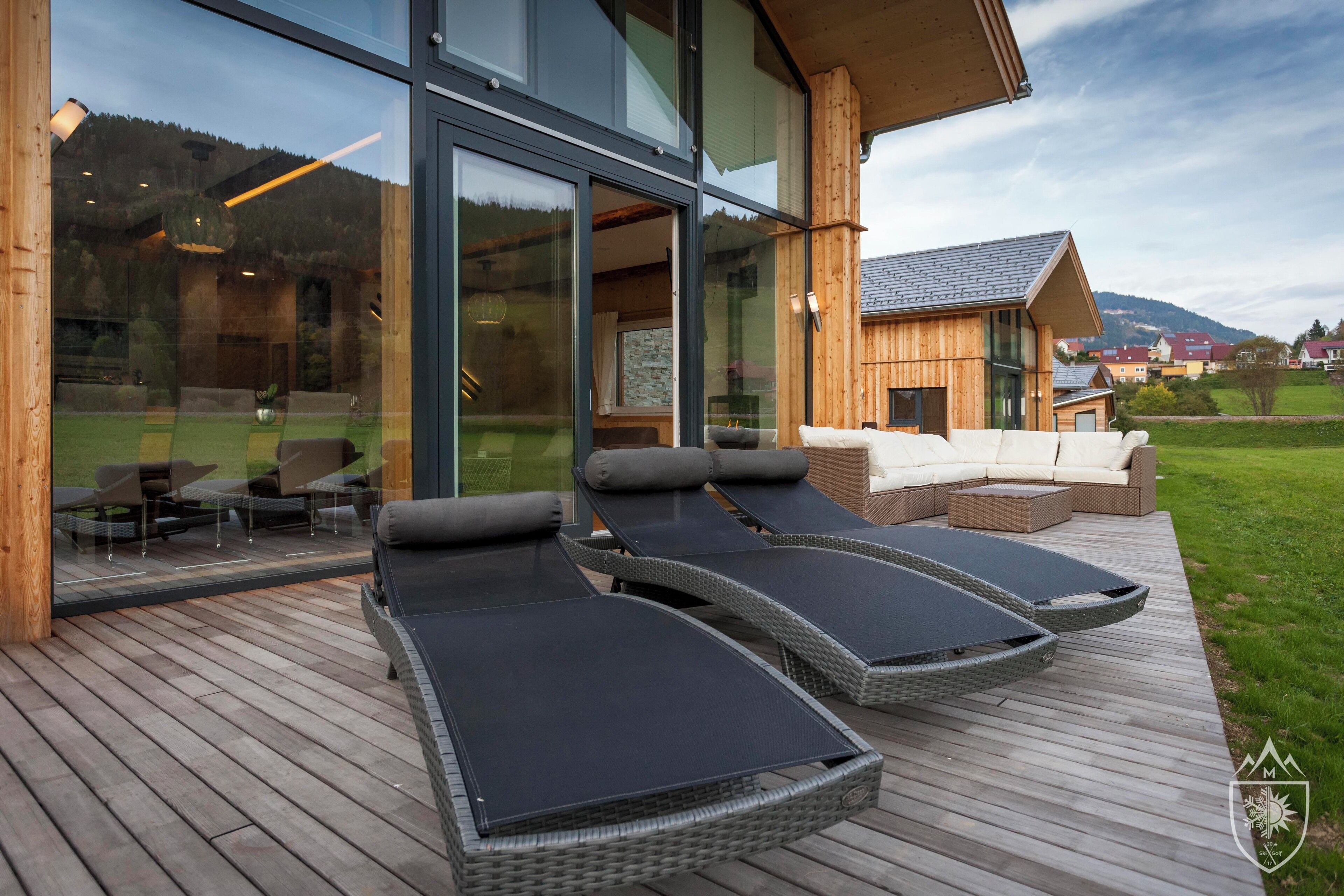 Une terrasse extérieure moderne, dotée de chaises longues et d’un canapé, de grandes fenêtres en verre et, en arrière-plan, d’une maison en bois.