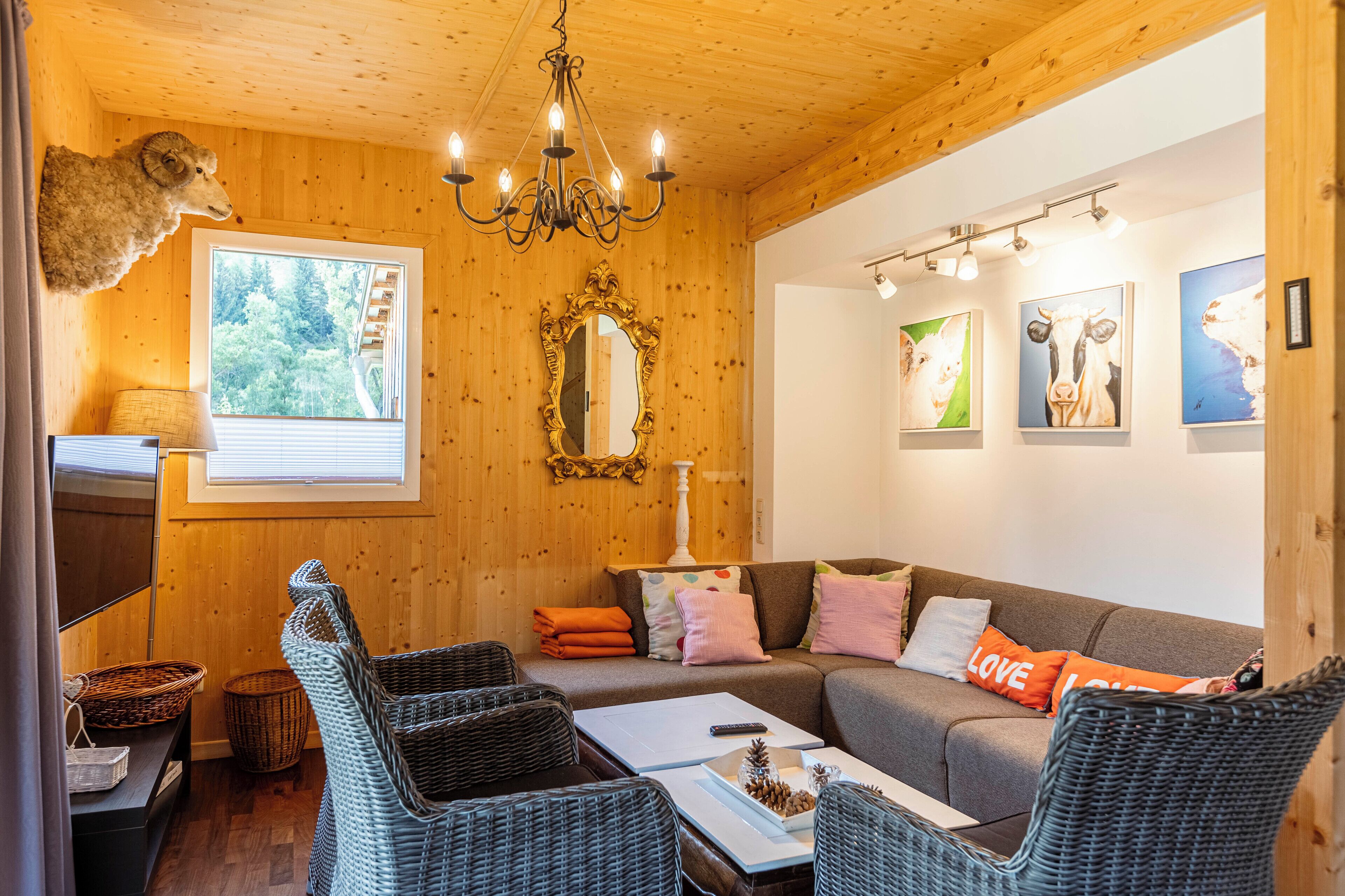 Een knus interieur met houten accenten, een kroonluchter, een bank met kleurrijke kussens, rieten stoelen en kunstwerken aan de muren.