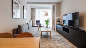 A modern living room with a sofa, a dining table, a TV on a black stand, and a large window with curtains. - King Apartamenty Galaxy (Szczecin)
