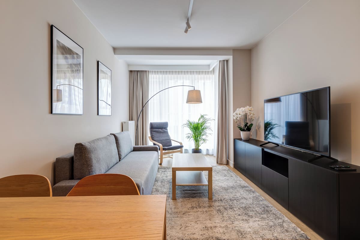 A modern living room with a sofa, a dining table, a TV on a black stand, and a large window with curtains.