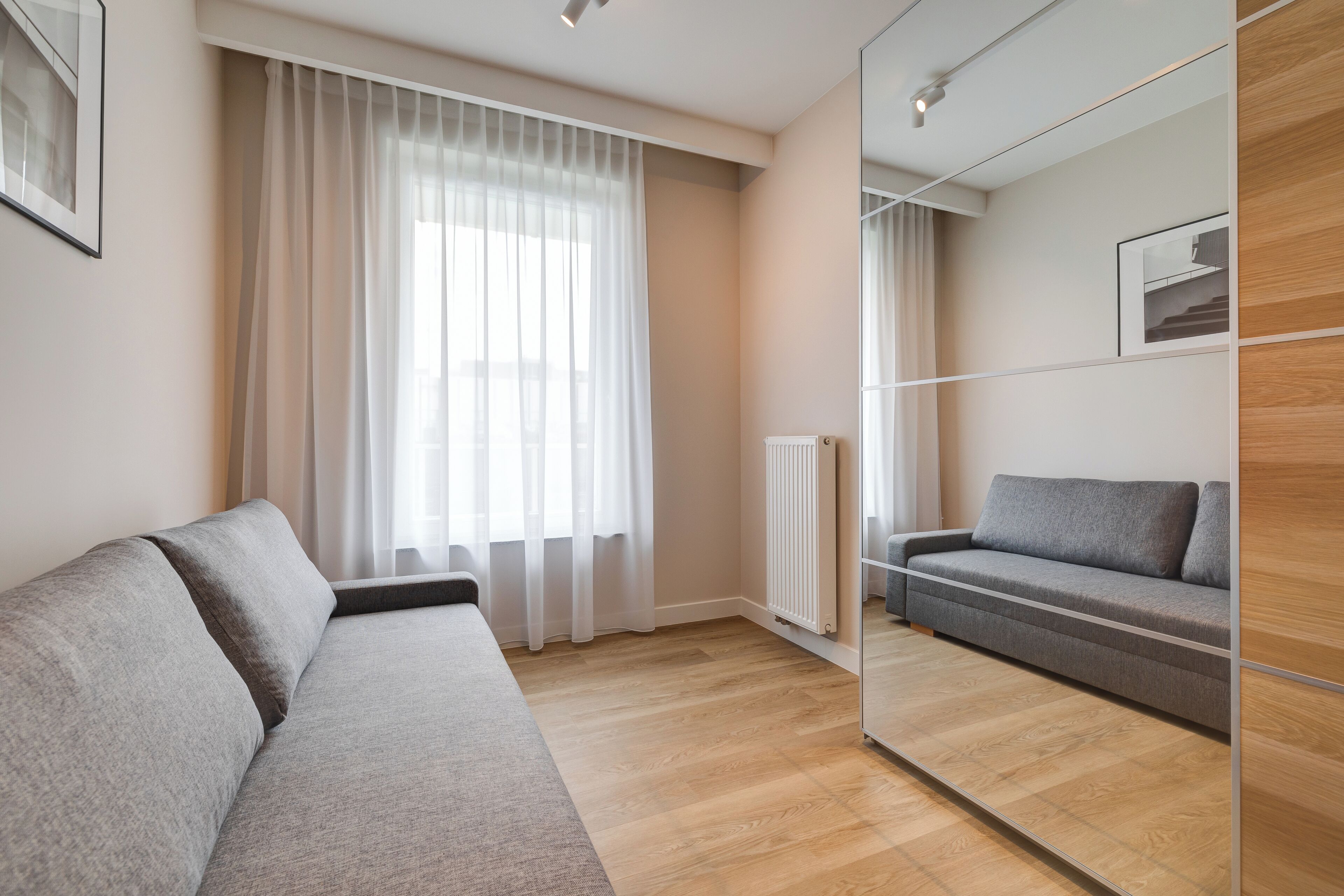 Een moderne woonkamer met een grijze bank, een houten vloer, een groot raam met dunne gordijnen en een spiegelwand.