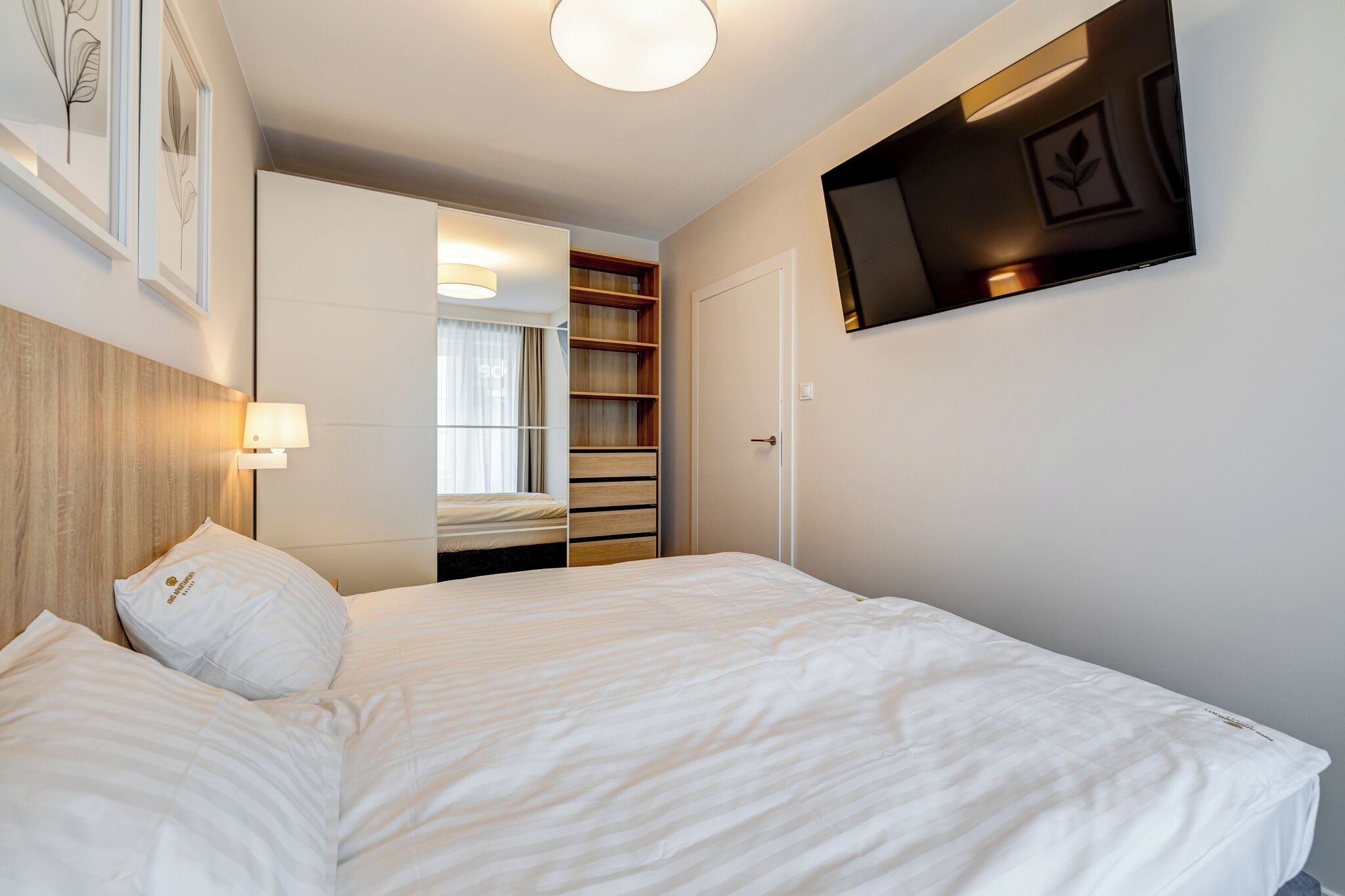 Une chambre d’hôtel comprenant un lit, une lampe de chevet, une armoire, un téléviseur fixé au mur et des œuvres d’art encadrées accrochées au mur.