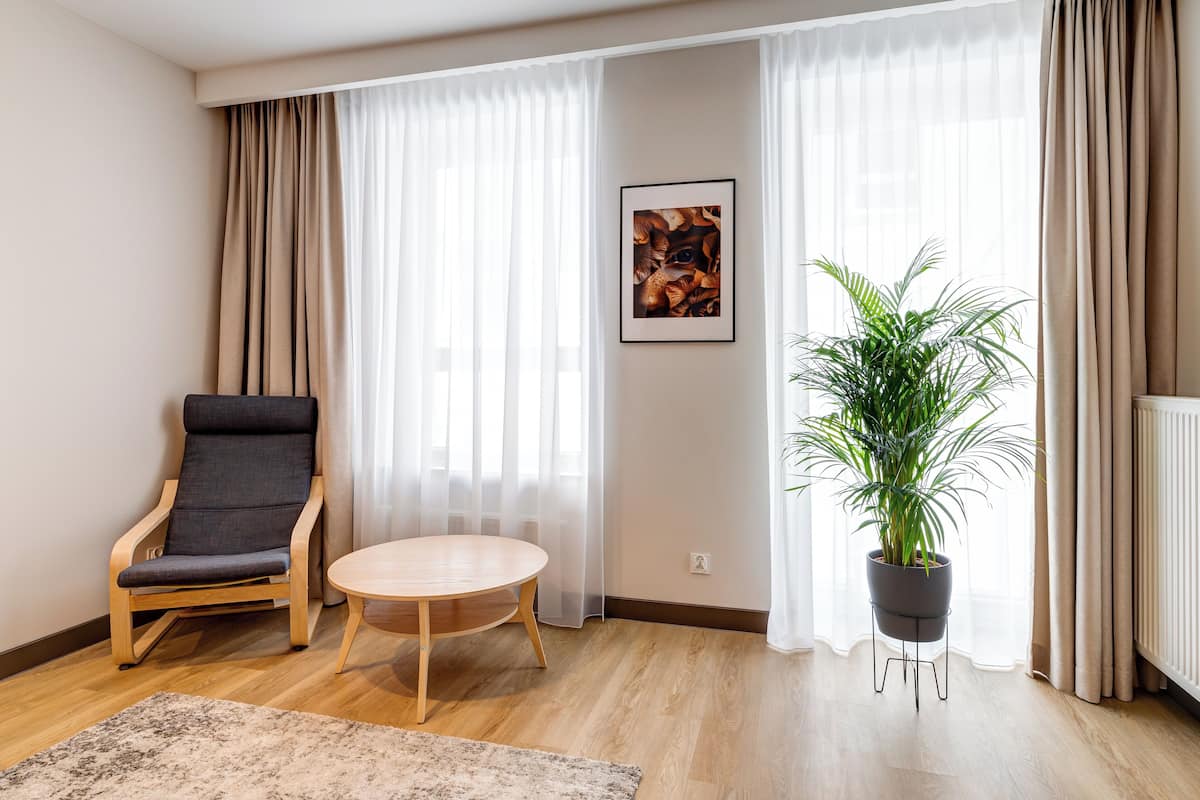 A modern living room with a wooden floor, a grey armchair, a round wooden table, a potted plant, and sheer curtains.