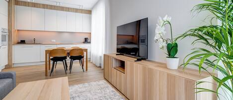 Uma cozinha moderna com armários brancos, um espaço de refeições com duas cadeiras, uma TV em um console de madeira e uma grande planta em vaso.