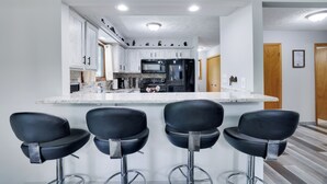 A modern kitchen with white cabinets, a black refrigerator, and a marble countertop. There are four black bar stools in front of the counter. - Hot Tub, Lake, and LOCATION! Fall Hiking, Adventures and Relaxation (Albrightsville)