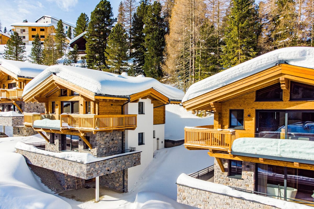 A snow-covered lodge with wooden balconies and stone foundations, surrounded by trees.