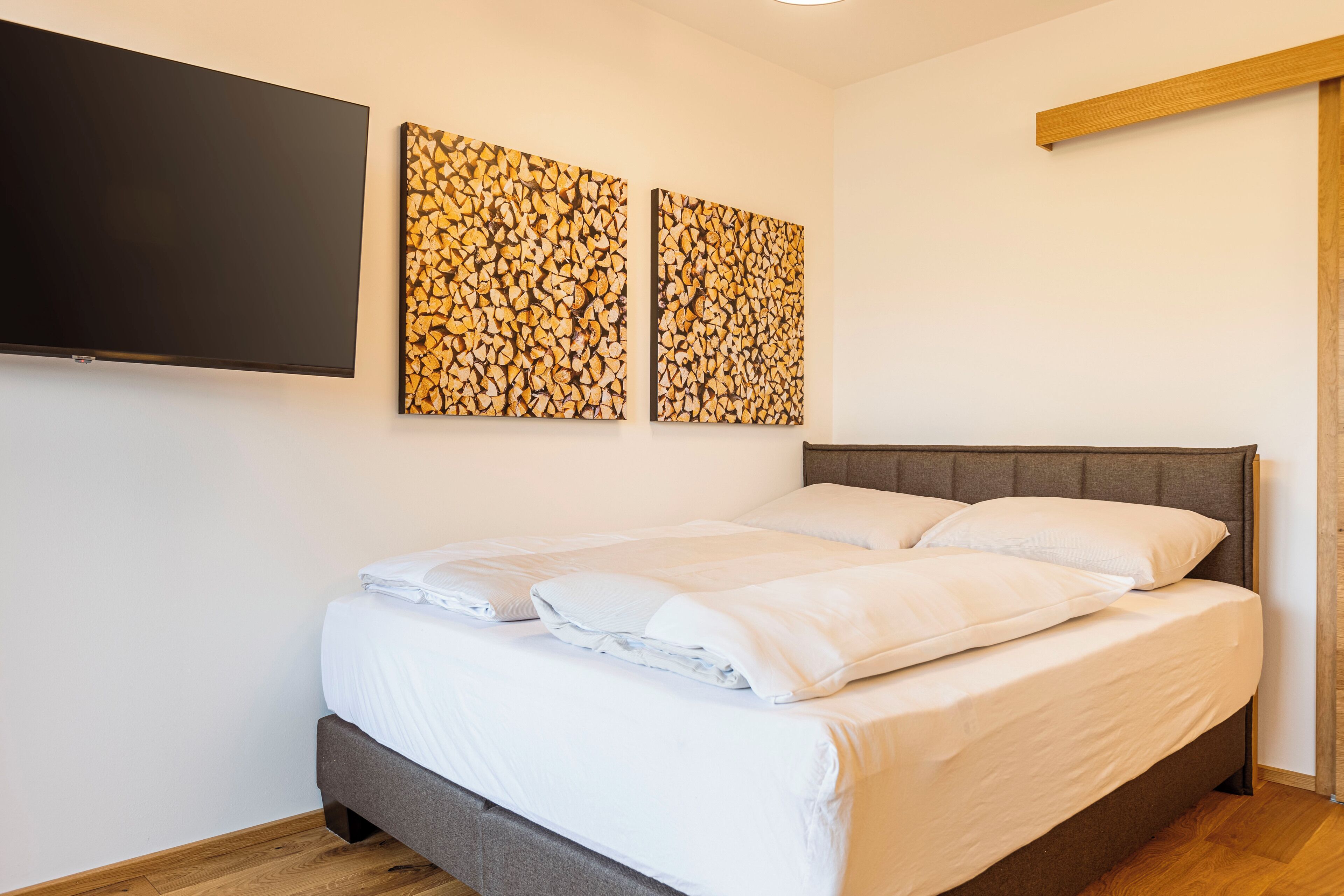 A neatly made bed with white linens and pillows, a flat-screen TV mounted on the wall, and two abstract art pieces above the bed.