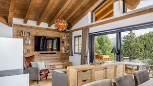 A modern living room with a wooden ceiling, a flat-screen TV, bookshelves, a sofa, and a dining area with a view of the outdoors. - Turrach Lodges by ALPS RESORTS (Stadl-Predlitz)
