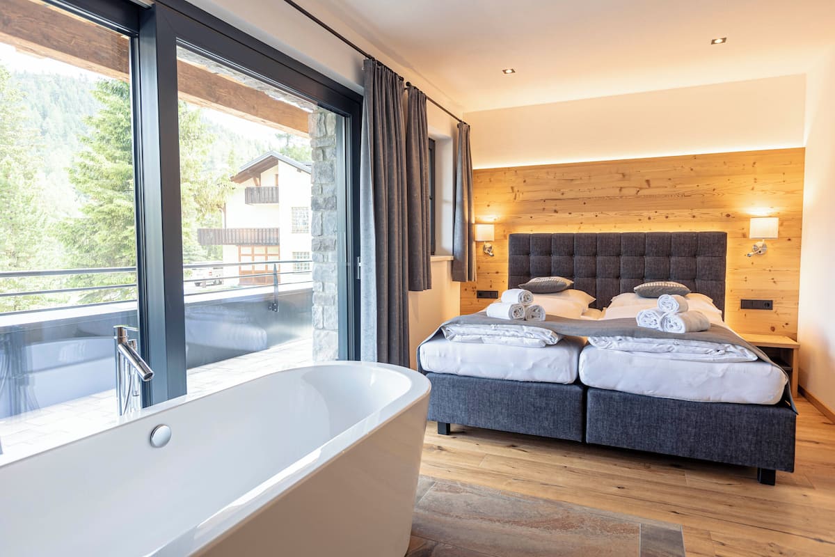 A modern bedroom with a large bathtub, a bed with neatly folded towels, and a view of a mountain cabin through large windows.