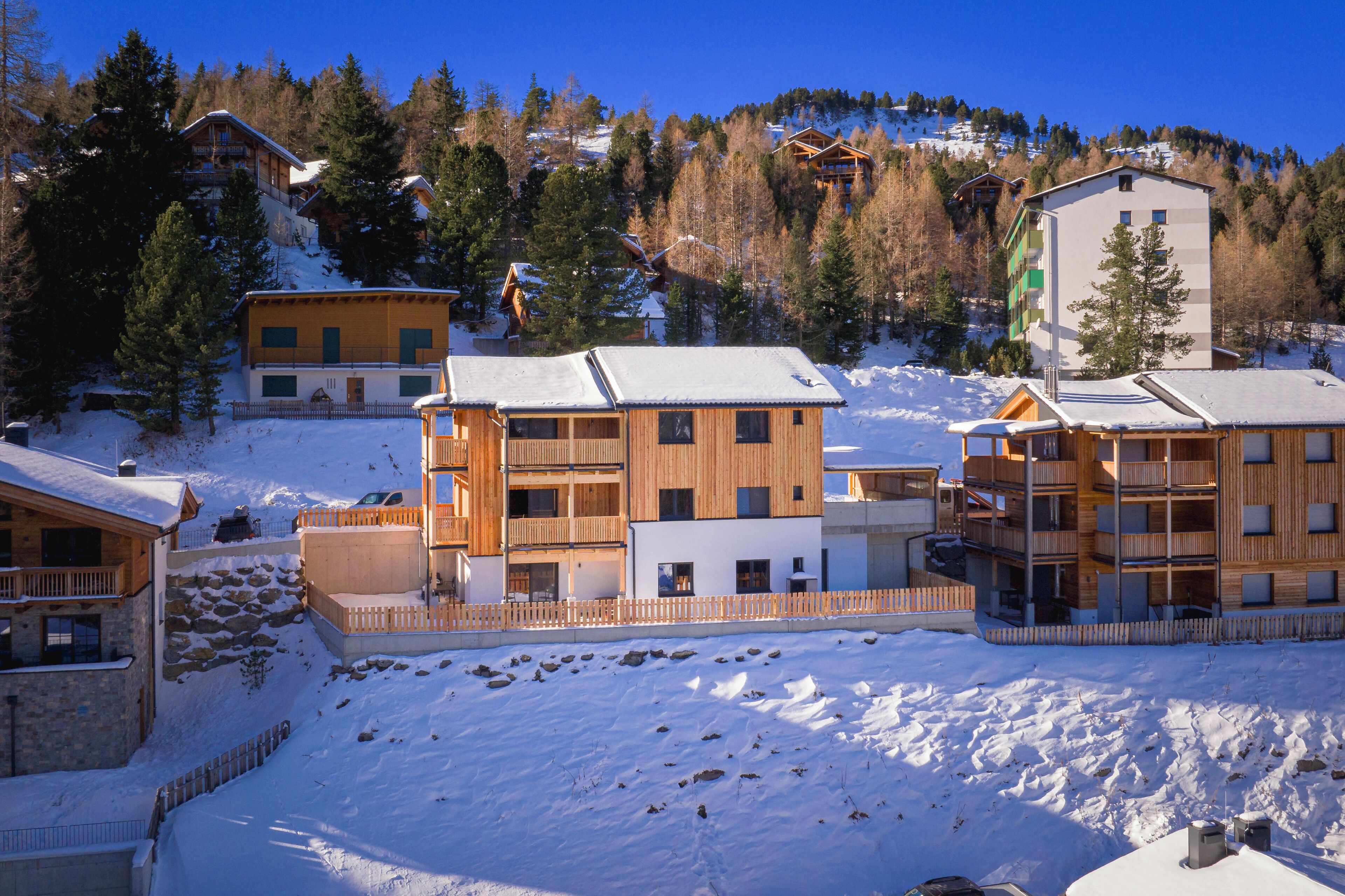 En snötäckt semesteranläggning med flera trähus, inklusive en stor byggnad i flera våningar och mindre stugor, omgiven av träd och med ett berg i bakgrunden.