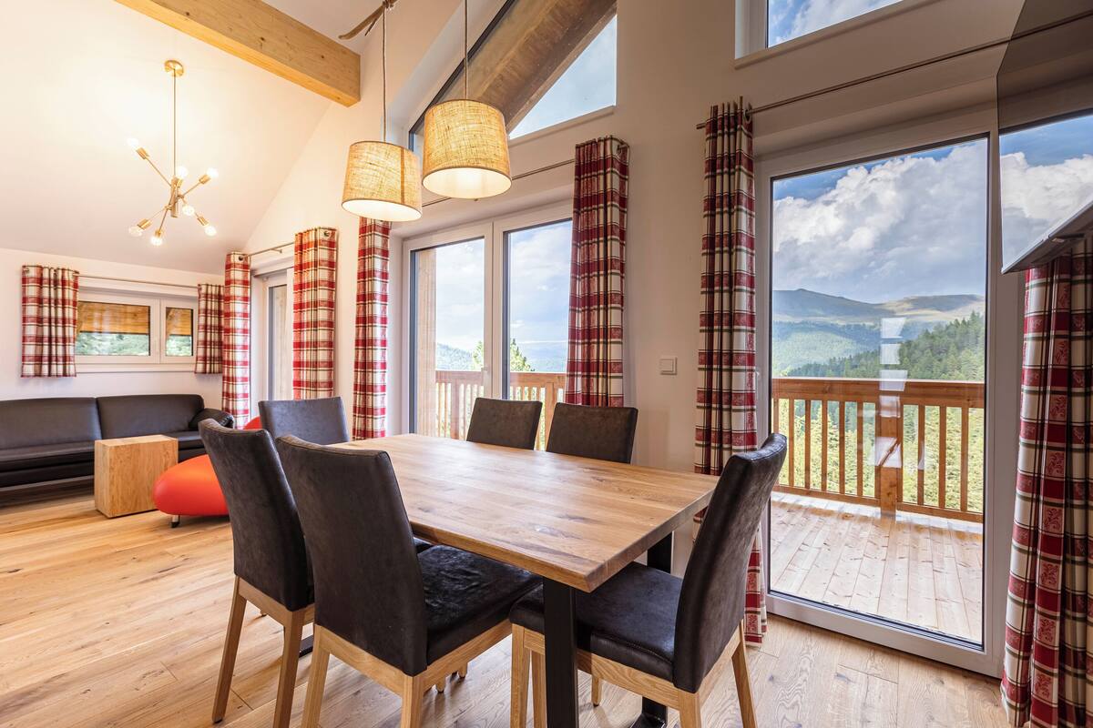 A dining area with a wooden table and chairs, large windows with plaid curtains, and a view of a wooden deck and mountains.