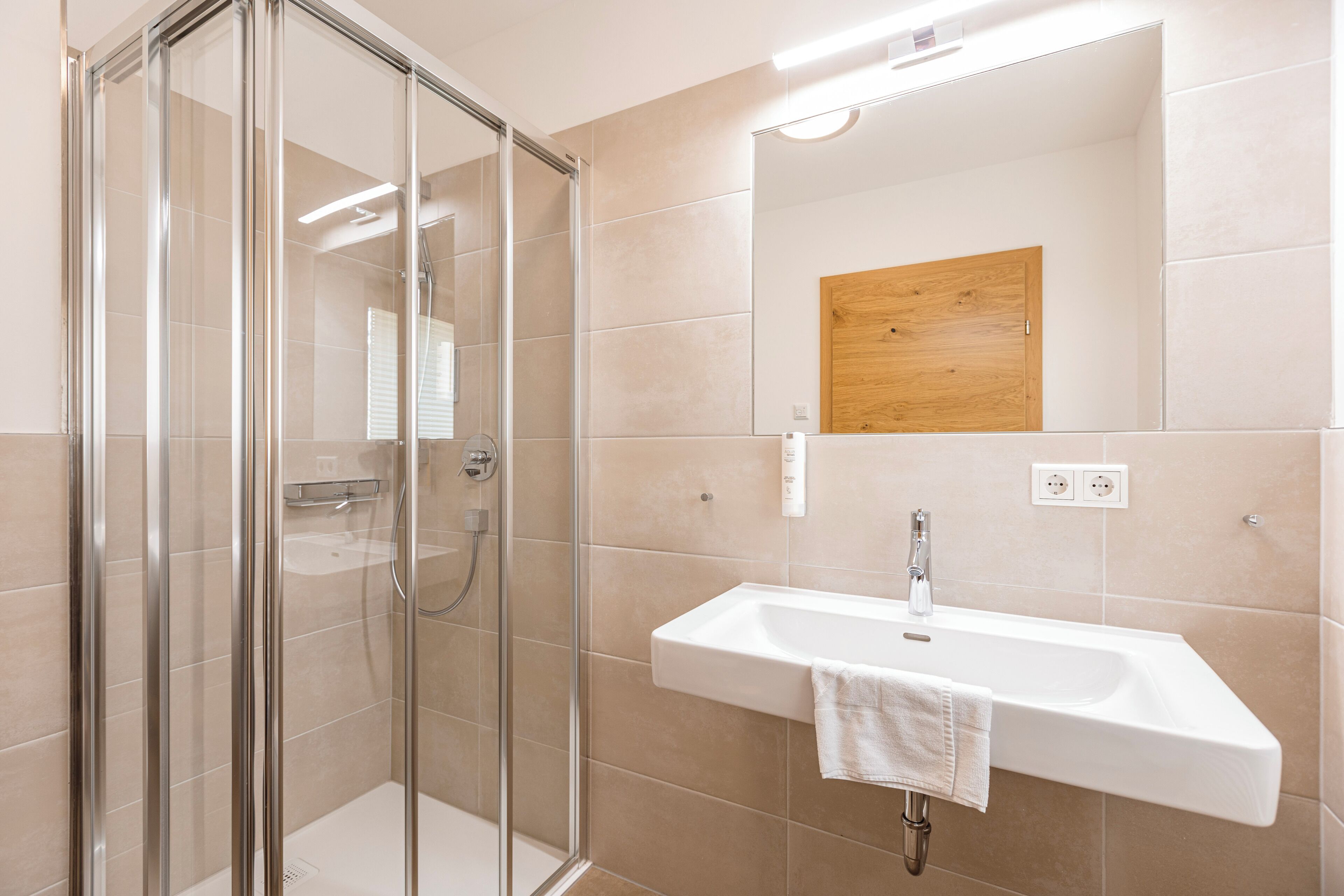 Un baño moderno con cabina de ducha de vidrio, un lavabo blanco y un panel de madera sobre el espejo.
