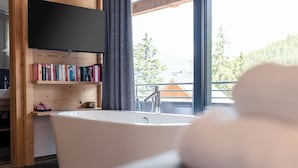 A modern bathroom with a freestanding bathtub, wooden shelves with books, and a view of a balcony with a railing. - Turrach Lodges by ALPS RESORTS (Stadl-Predlitz)