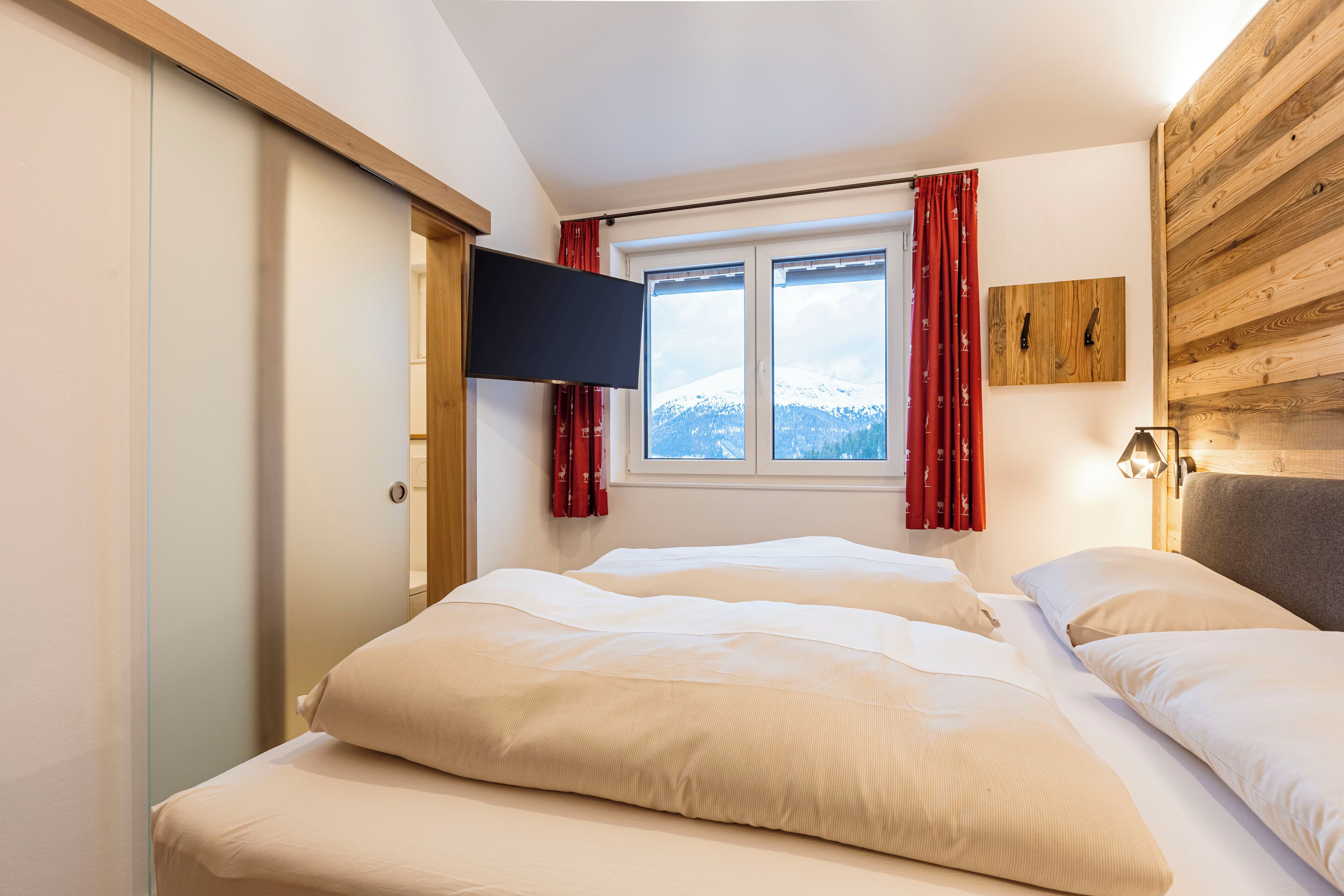 A bedroom with two beds, a TV mounted on the wall, a window with red curtains, and a view of snow-capped mountains.