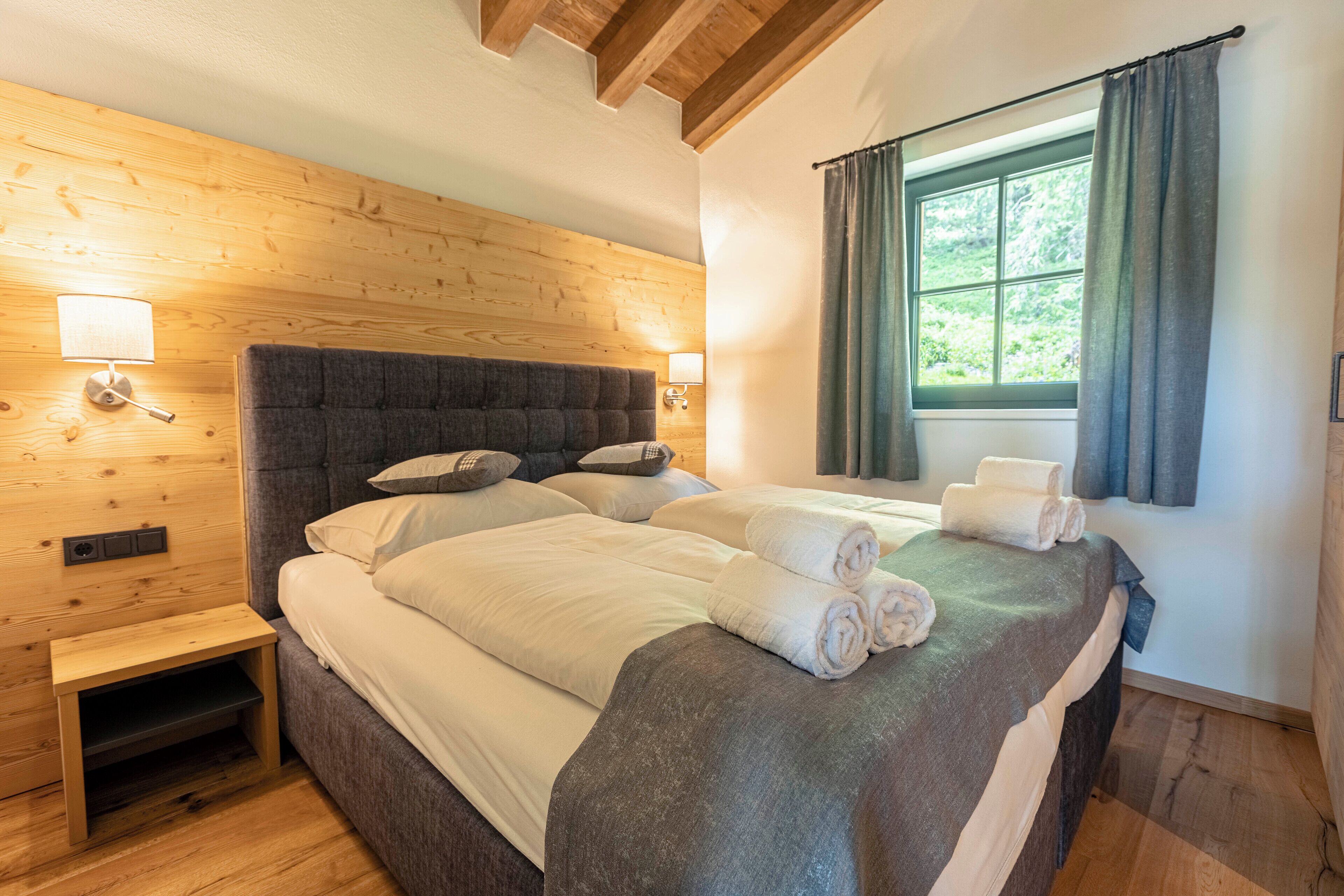Une chambre à coucher avec une tête de lit en bois, deux lits avec du linge de lit blanc, une table de chevet, une commode et une fenêtre avec des rideaux.