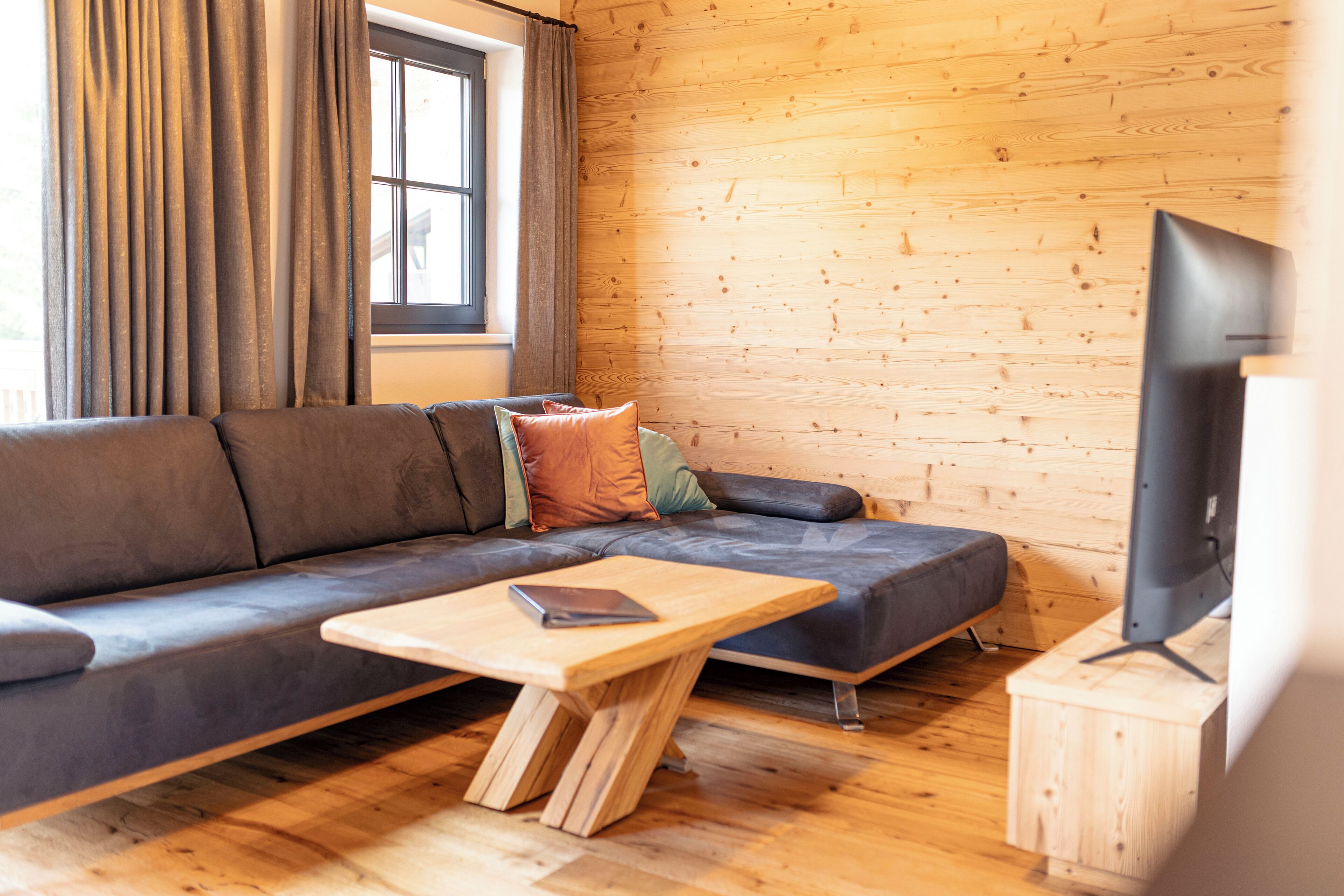 A modern living room with a wooden floor, a grey sofa with cushions, a wooden coffee table, and a television on a wooden stand.