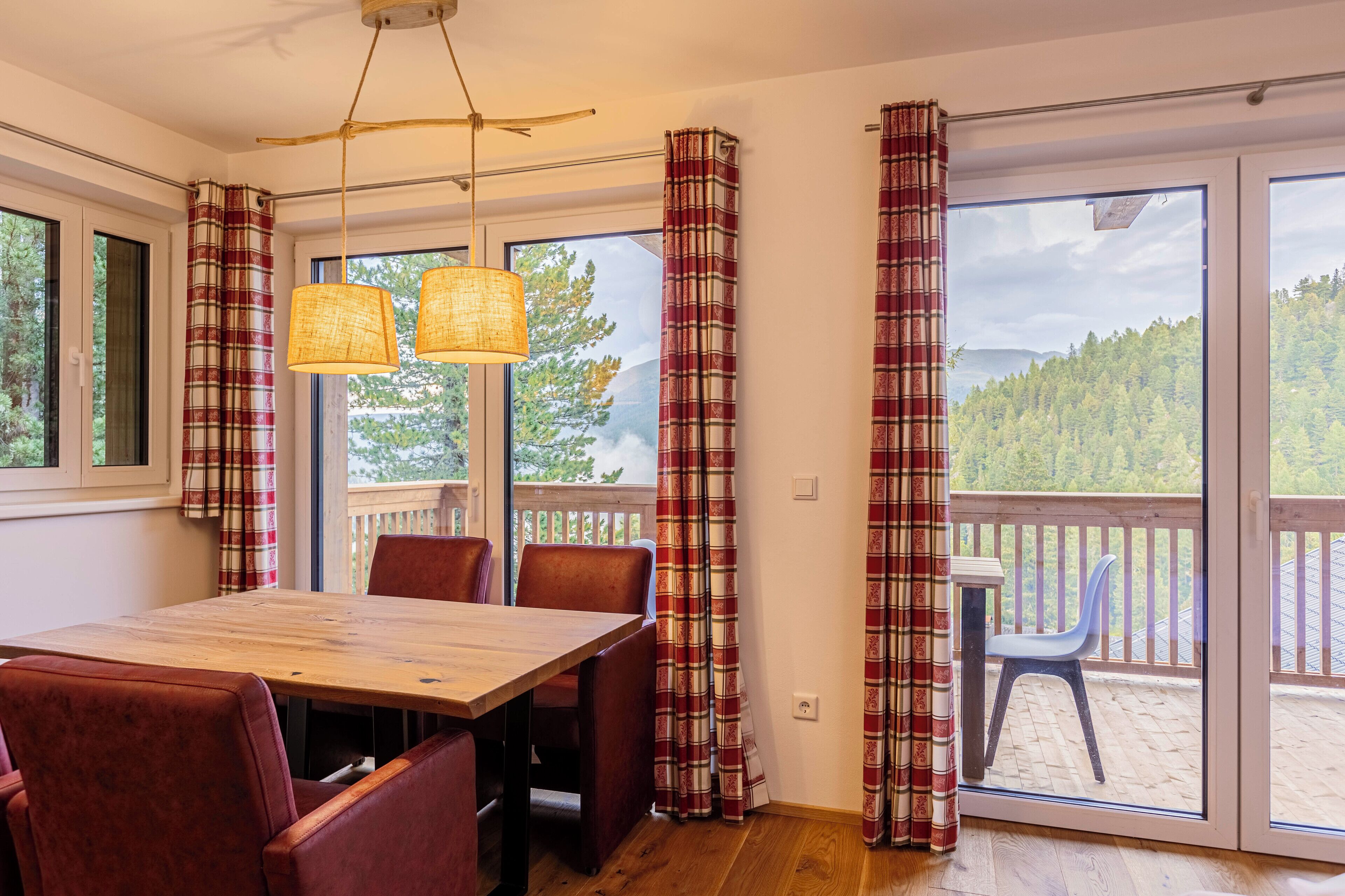 Un comedor con mesa y sillas de madera, ventanas amplias con cortinas, y vista a una terraza y bosque.