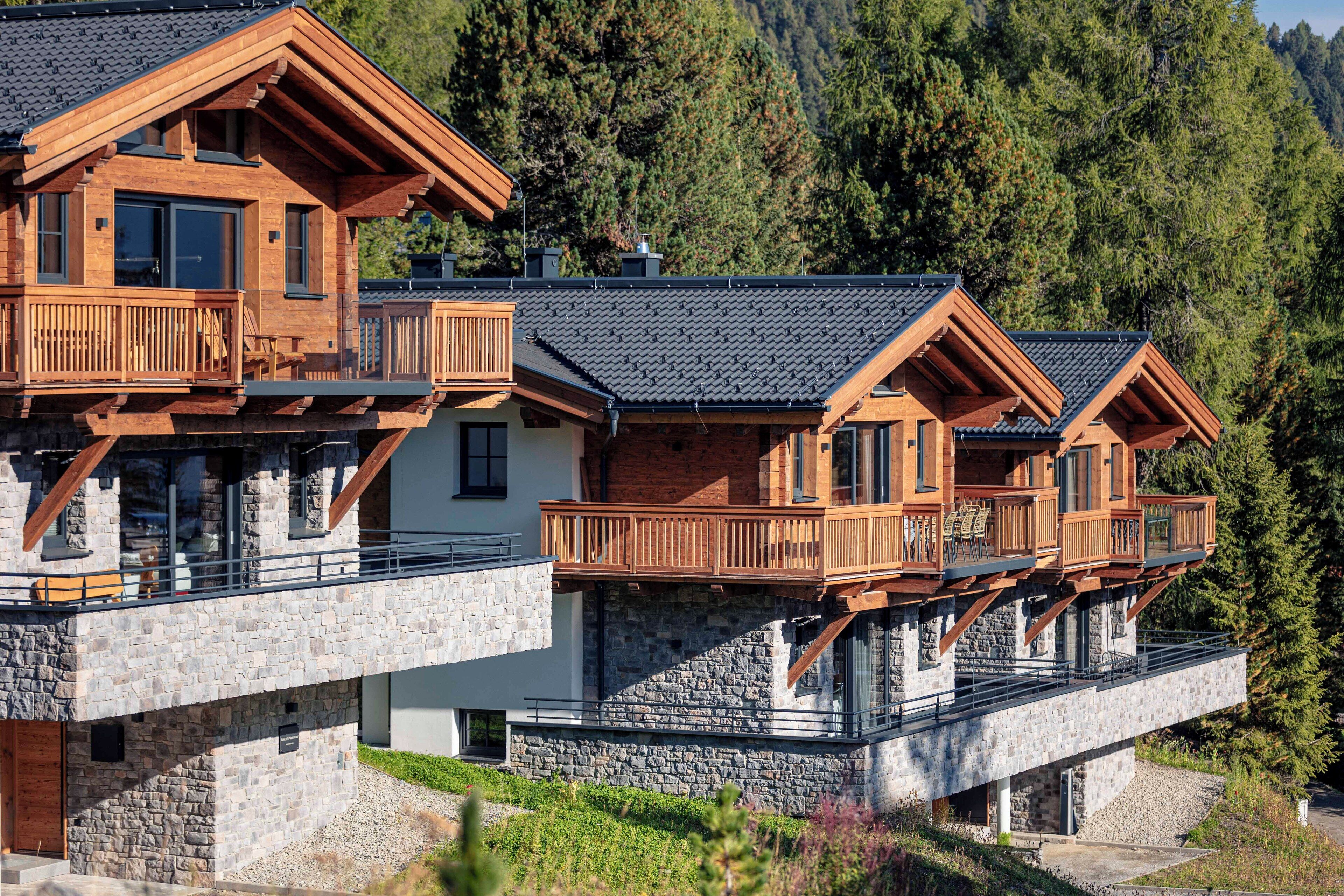 Una cabaña de madera de varios pisos con balcones, cimientos de piedra y un paisaje montañoso de fondo.