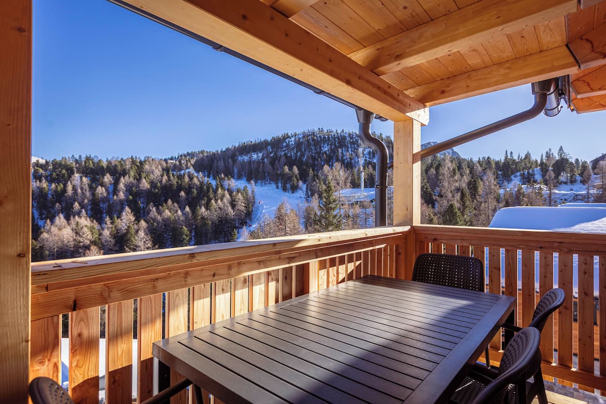 A wooden deck with a table and chairs, overlooking a snowy landscape with trees and a mountain.